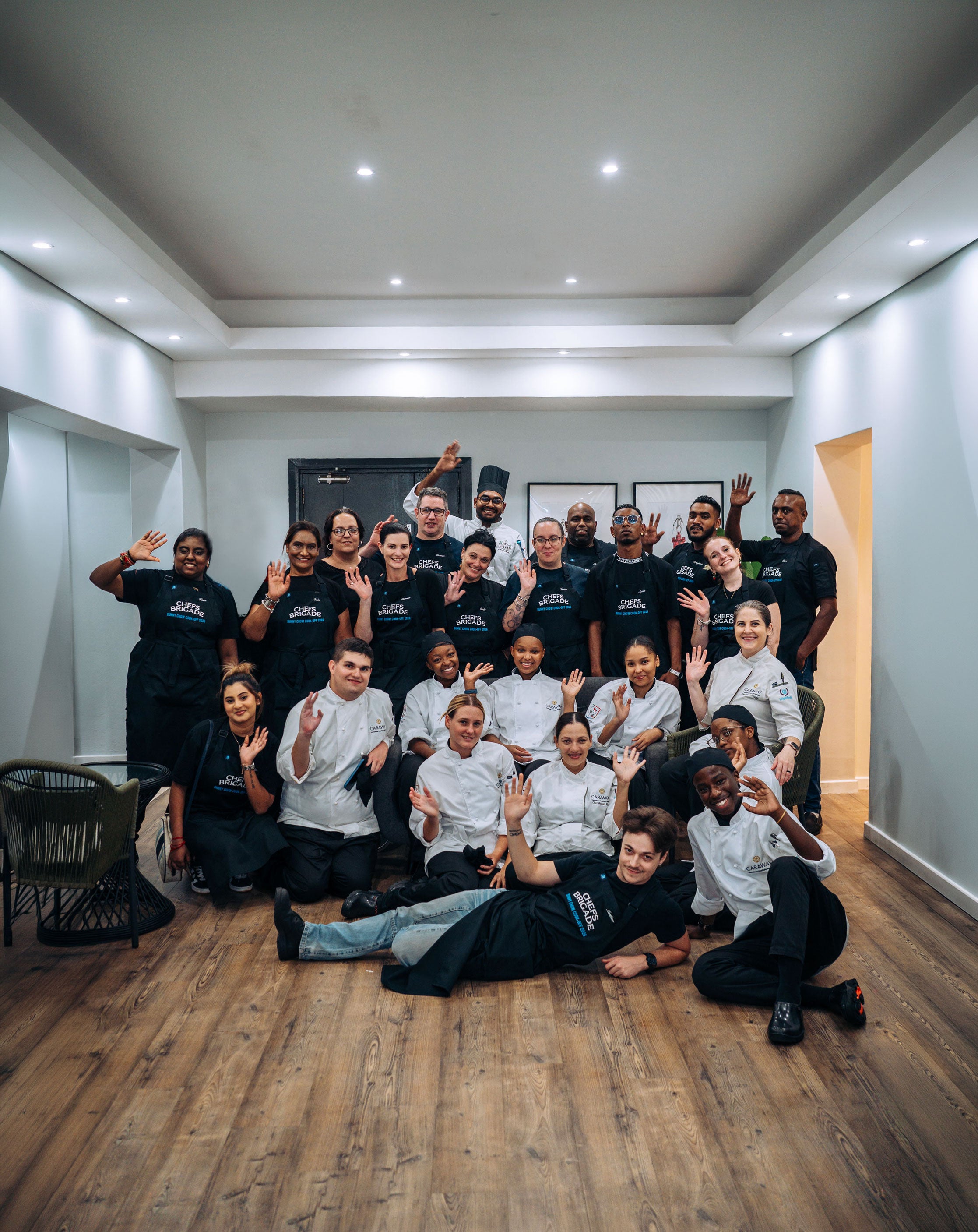 Group of people in black and white chef uniforms posing together in a room with wooden flooring and white walls.