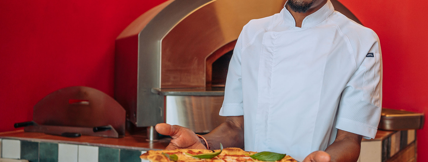 Chef holding a pizza in front of a pizza oven with a red wall background