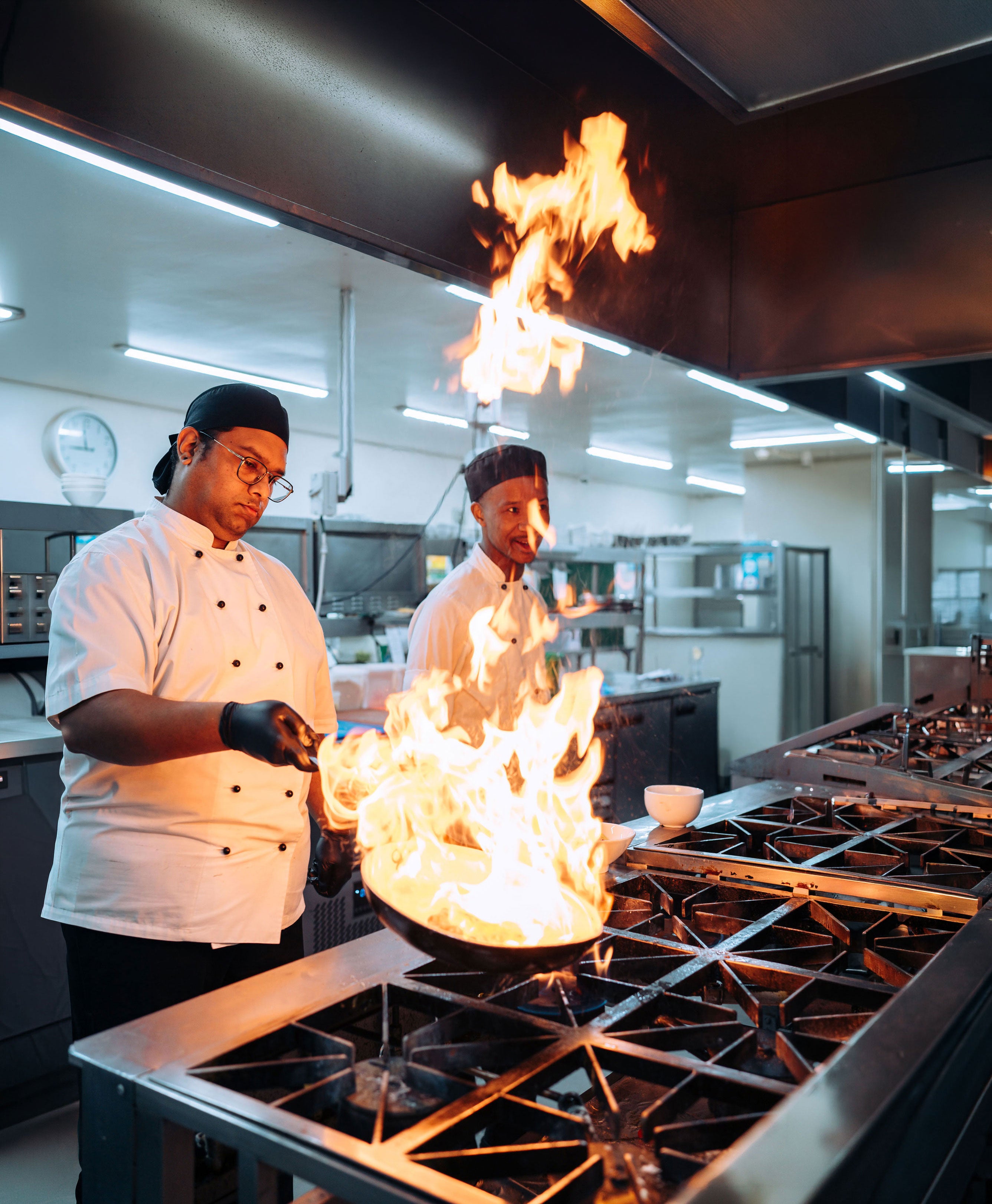 Two chefs in a kitchen with flames coming from a pan on a stove.