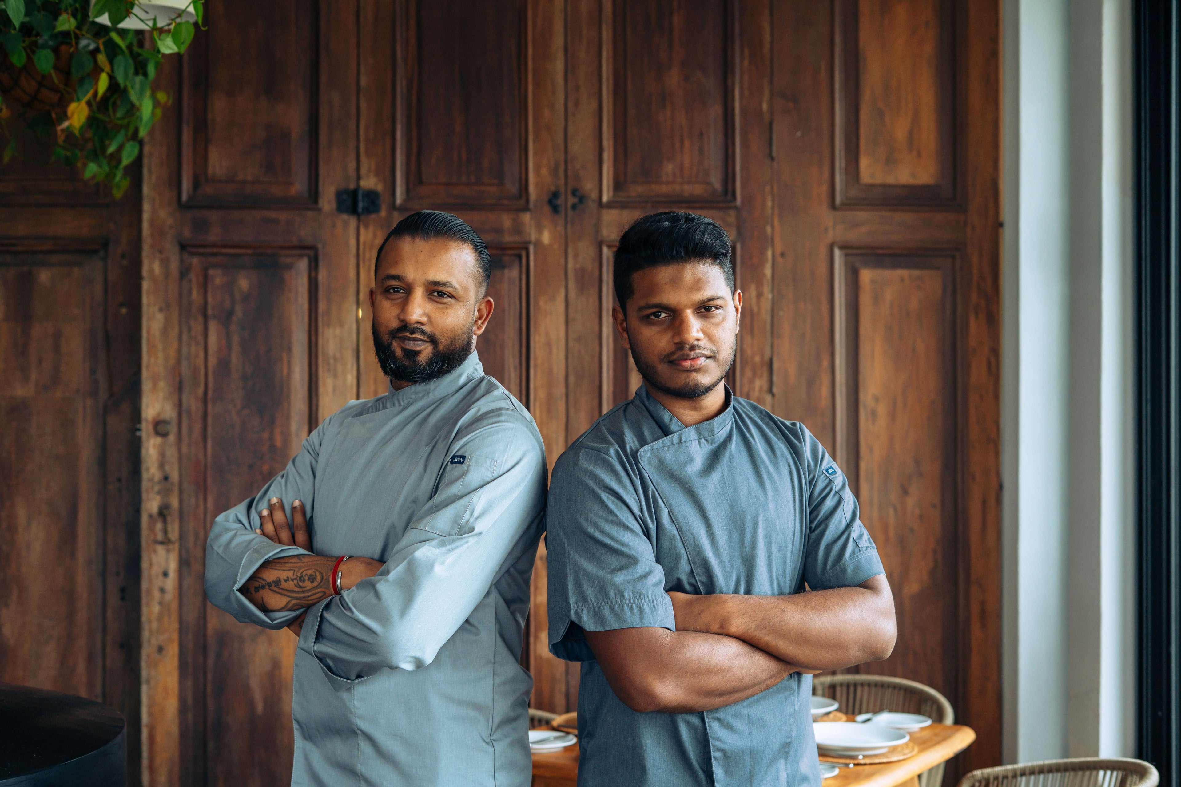Two men in gray chef coats standing in a kitchen with wooden cabinets.