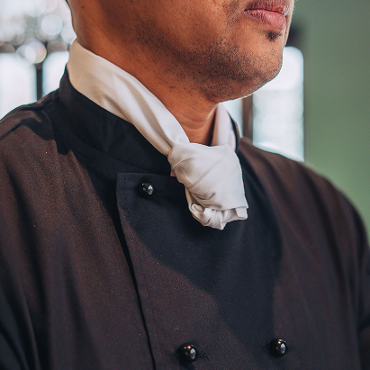 Close-up of a person wearing a black chef's coat with a white necktie