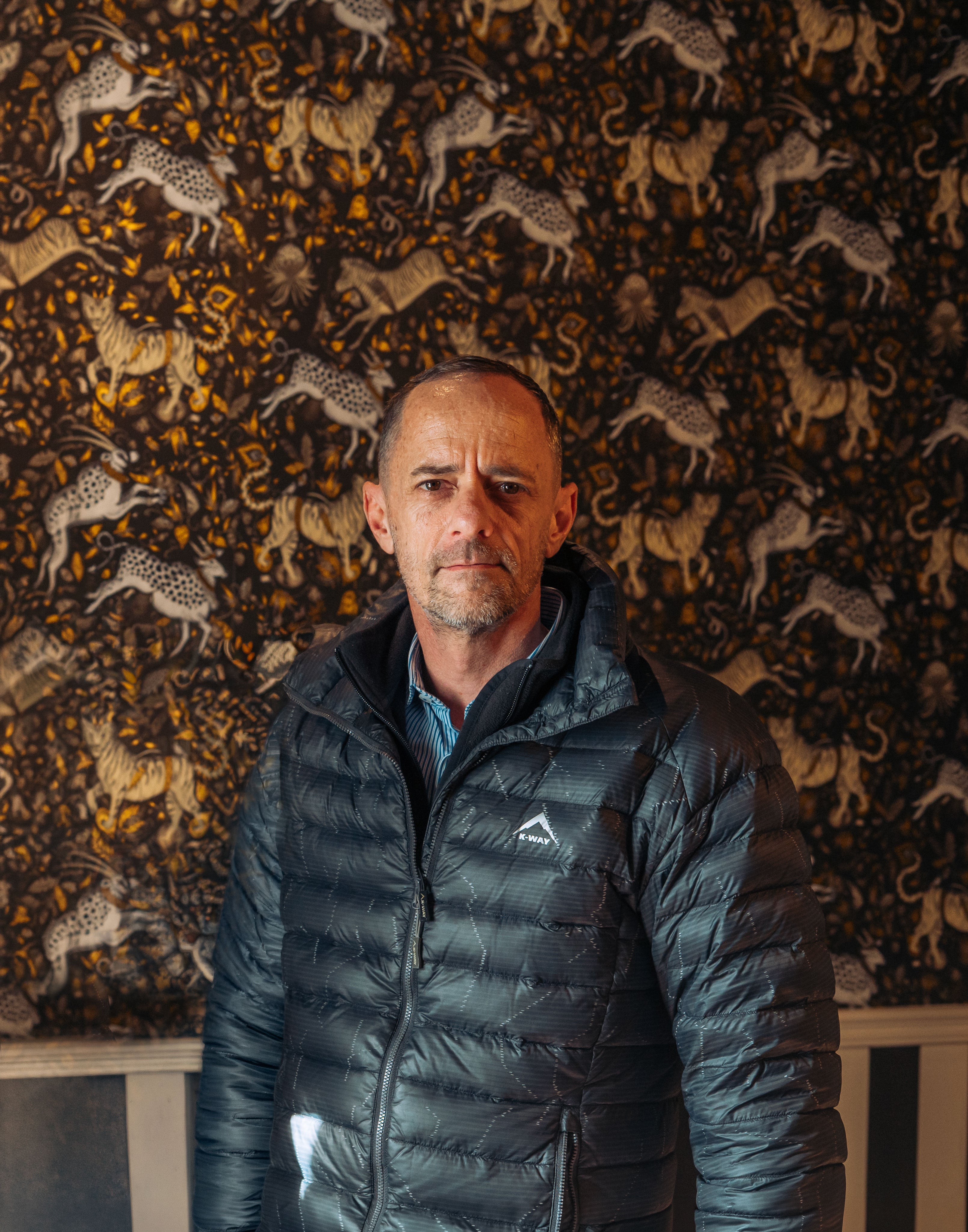Man wearing a dark blue puffer jacket with a logo in front of a patterned wall.