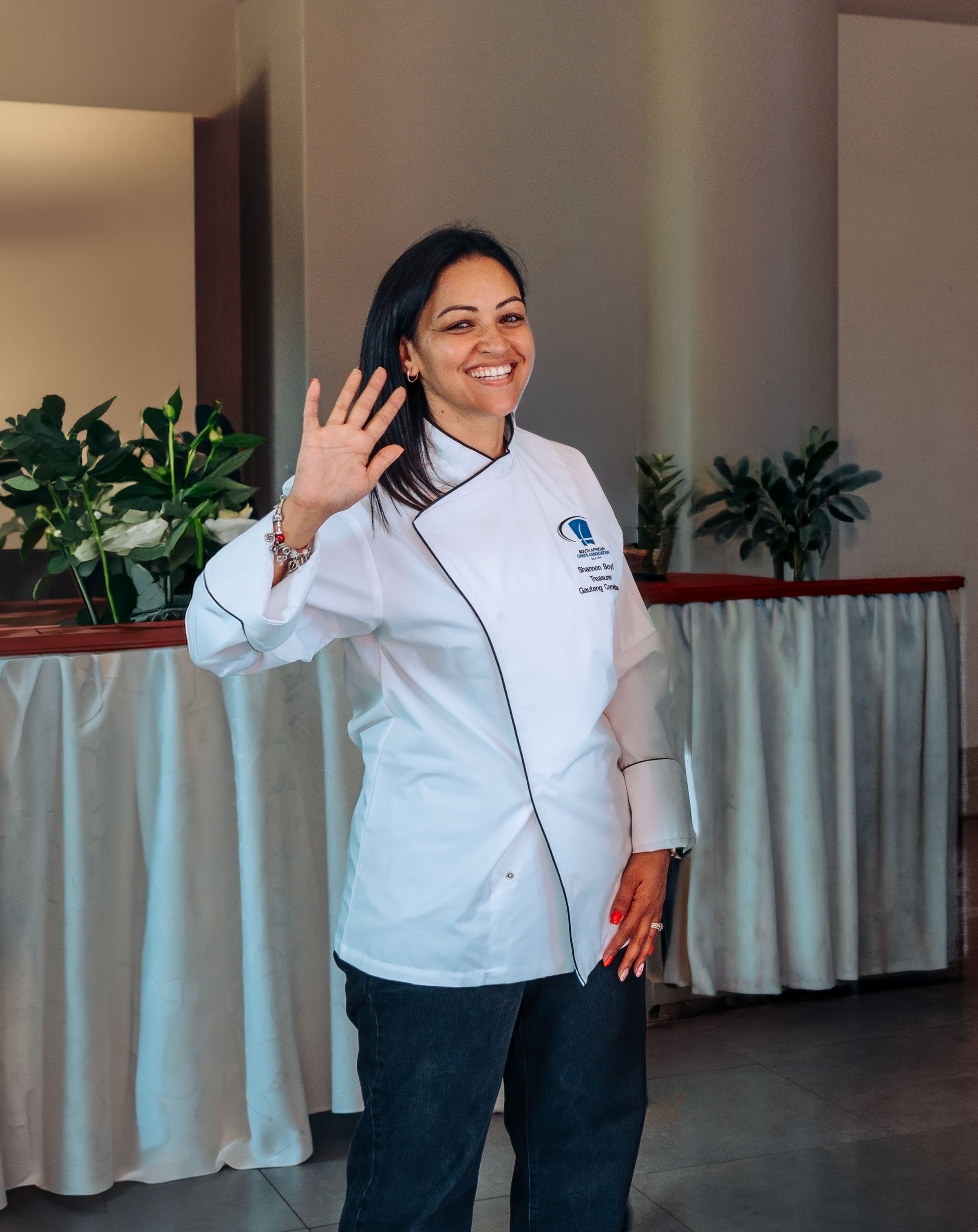Woman in a white chef's coat waving at the camera with plants in the background.