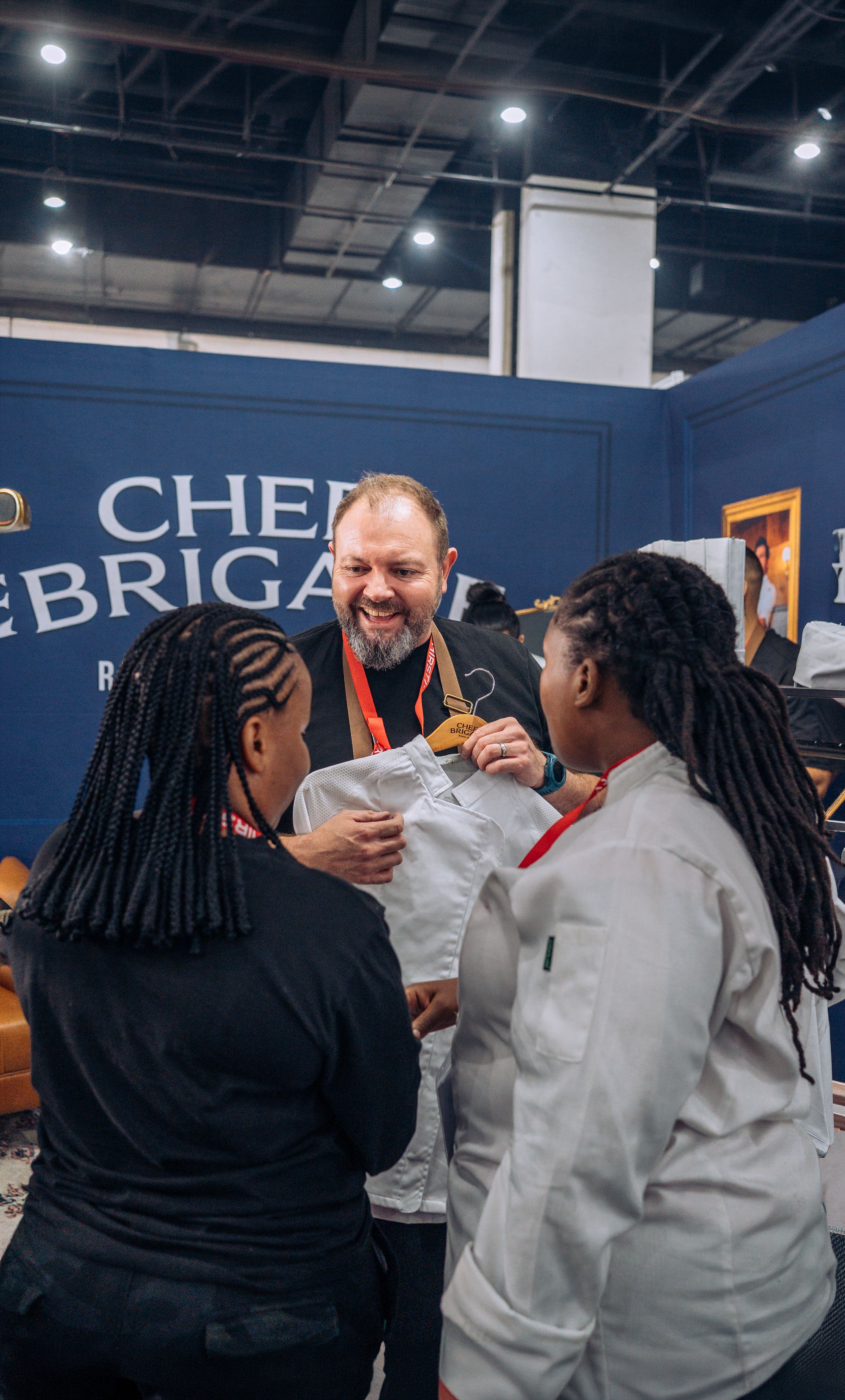 Man showing a white shirt to two women in front of a blue backdrop with 'Chefs Brigade' text.