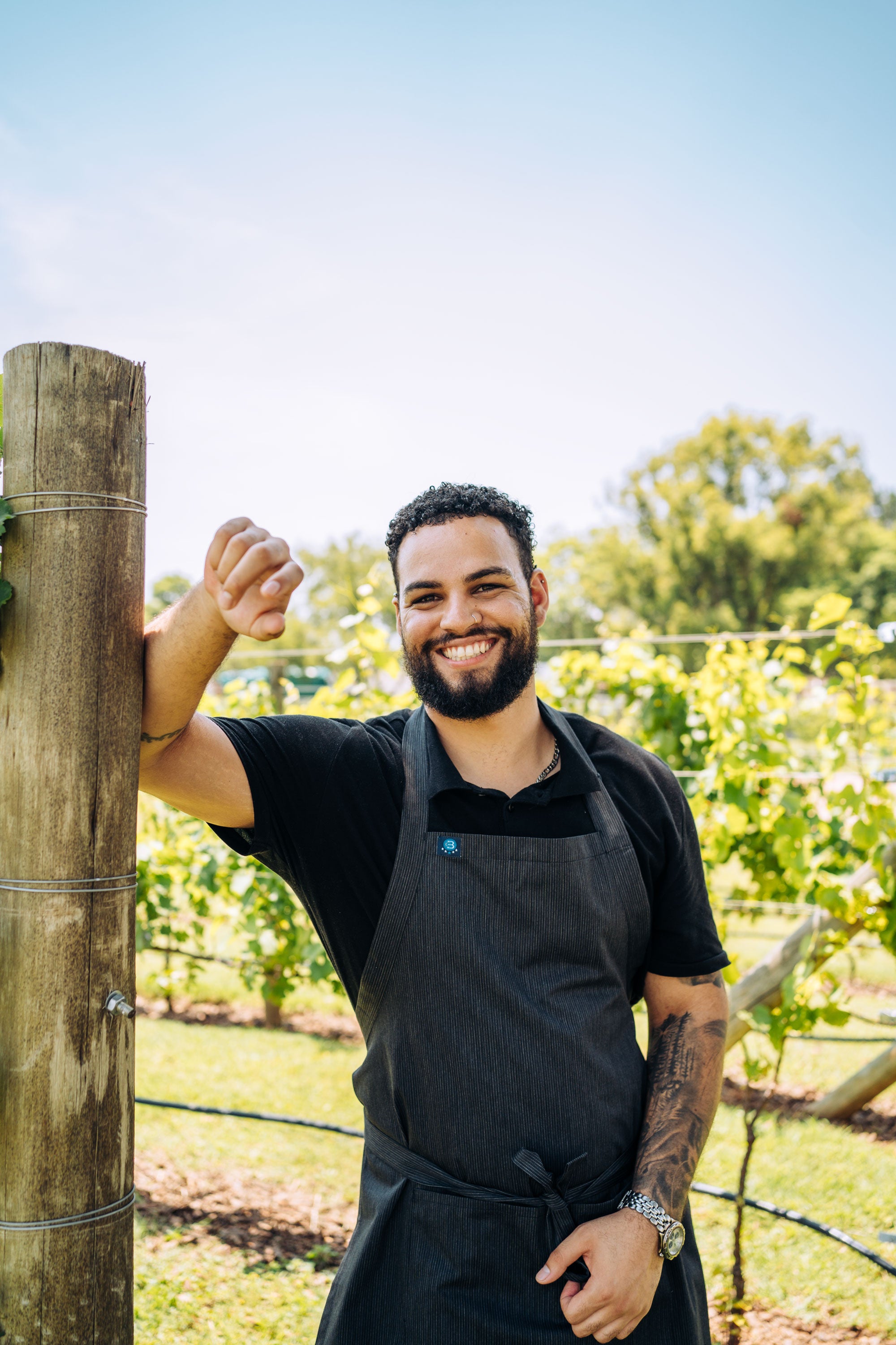 Man in a black apron standing in a vineyard, smiling.