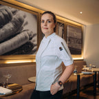 A female chef in a white uniform standing in a restaurant setting with framed wall art and tables in the background.