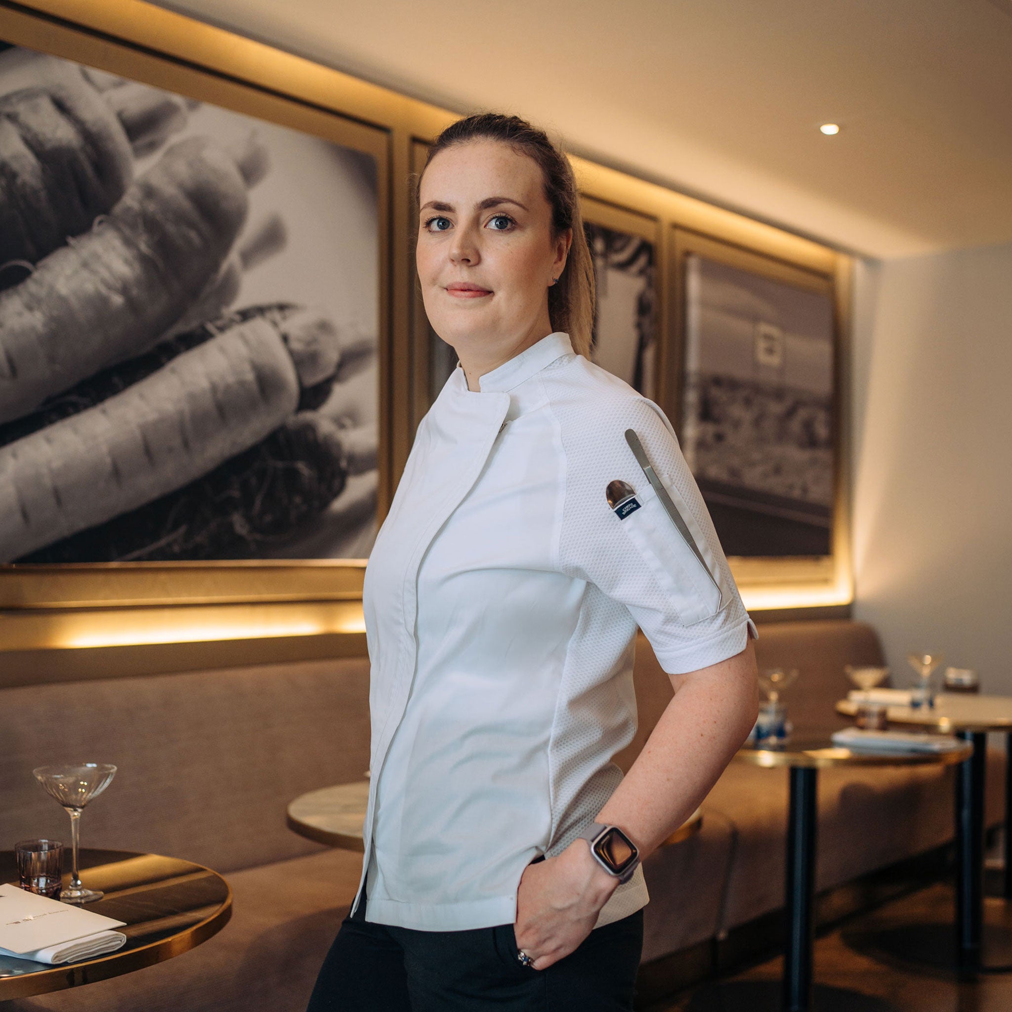 A female chef in a white uniform standing in a restaurant setting with framed wall art and tables in the background.