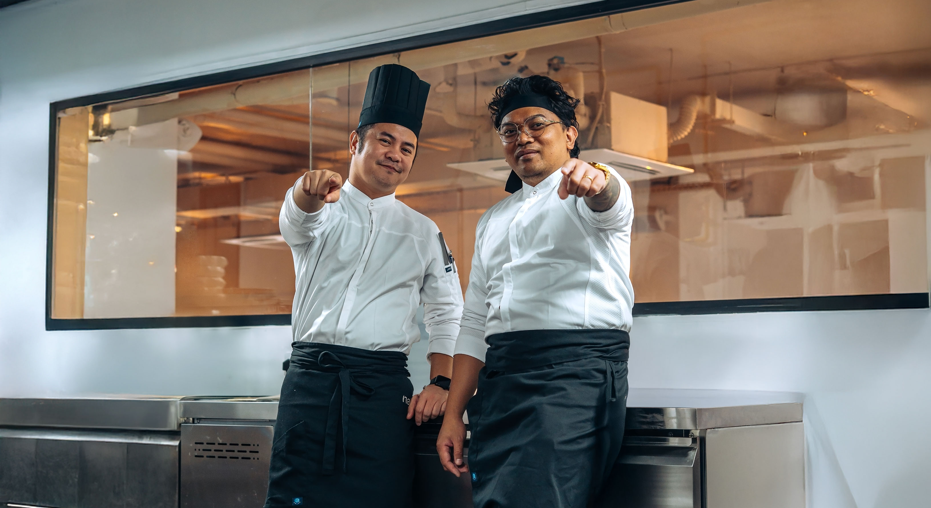 Two chefs in a professional kitchen pointing towards the camera.